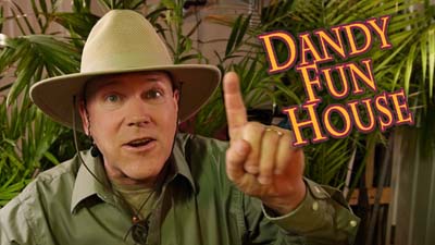 neil dandy wearing jungle explorer outfit in front of foliage next to dandy fun house logo pointing up with one finger