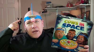 neil dandy wearing headband with flying saucer attached and toy cow magnetically attached to flying saucer while hold the box for cows in space